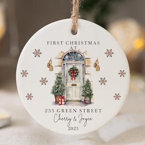 May include: A circular Christmas ornament featuring a watercolour illustration of a front door, Christmas trees, and a gift. Text reads "First Christmas At 235 Green Street Cherry & Joyce 2025". The ornament is suspended by a natural twine string.