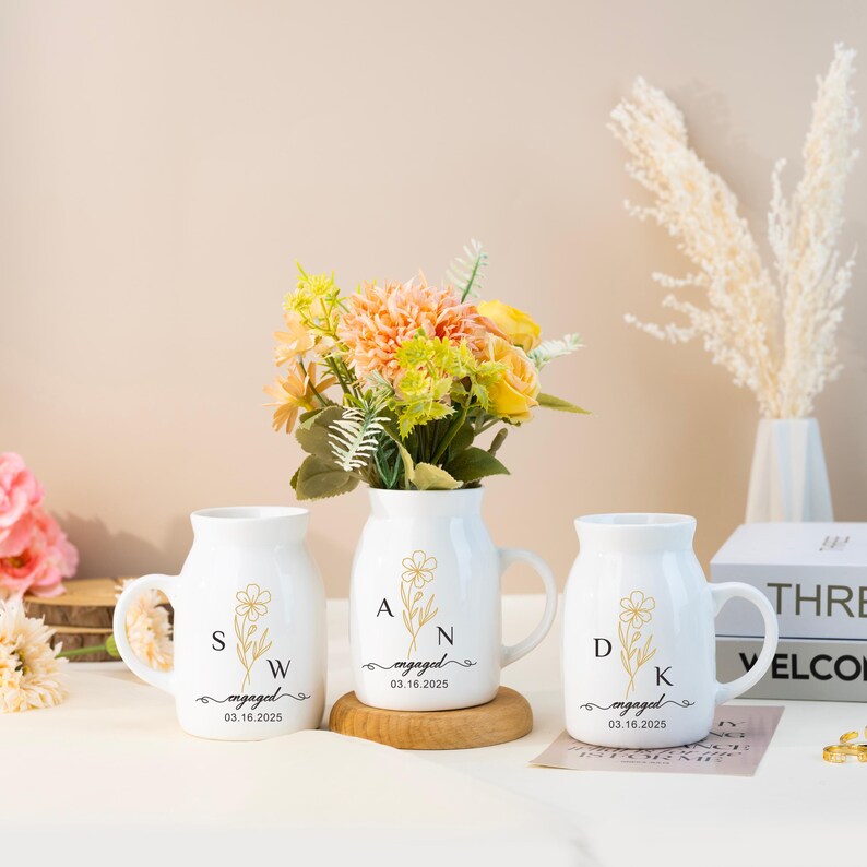 May include: Three white ceramic mugs with gold lettering and floral designs. The mugs say "engaged" with the date "03.16.2025" and initials "SW", "AN", and "DK".