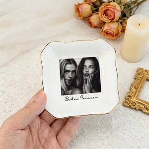 K&ouml;nnte beinhalten: Wei&szlig;es Keramik-Schmucksch&auml;lchen mit goldenem Rand, das ein Schwarz-Wei&szlig;-Foto von zwei Frauen mit dem Text "Bestie Forever" zeigt. Das Sch&auml;lchen hat einen gewellten Rand und wird von einer Hand gehalten. Dekorative Elemente befinden sich im Hintergrund.