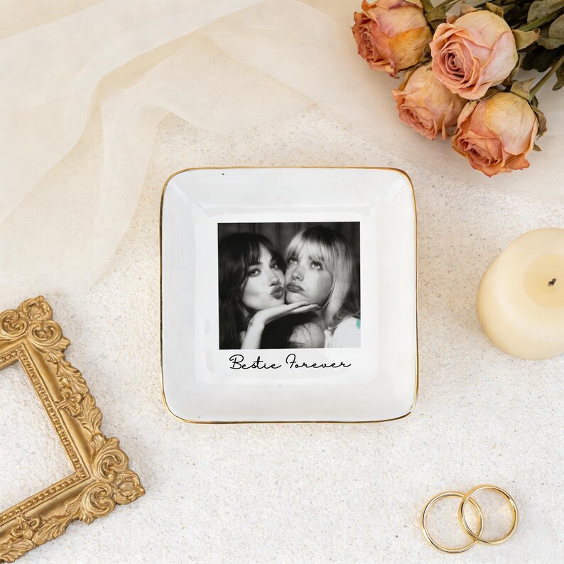 K&ouml;nnte beinhalten: Eine wei&szlig;e quadratische Schmuckschale mit goldenem Rand zeigt ein Schwarz-Wei&szlig;-Foto von zwei sich k&uuml;ssenden Frauen. Die Worte "Bestie Forever" sind unter dem Foto in Schreibschrift geschrieben. Die Schale ist von Blumen und einer Kerze umgeben.