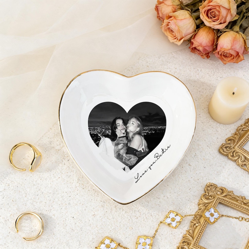 K&ouml;nnte beinhalten: Eine herzf&ouml;rmige wei&szlig;e Keramik-Schmuckschale mit goldenem Rand. Die Schale zeigt ein Schwarz-Wei&szlig;-Foto von zwei Personen und den handgeschriebenen Text "Love you Bestie". Goldene Ringe, eine Kerze und Blumen sind ebenfalls vorhanden.