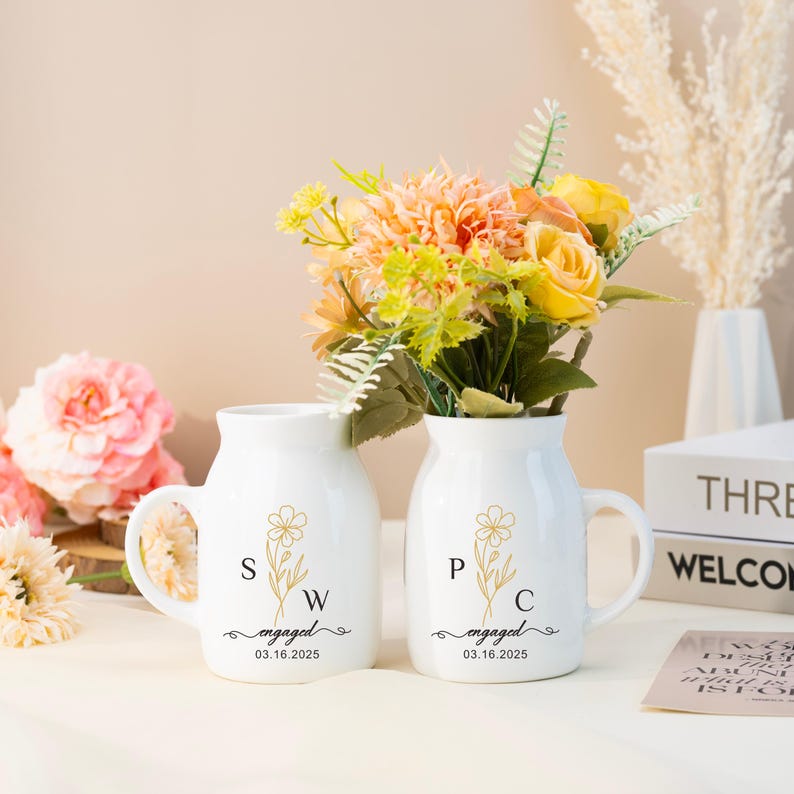 May include: Two white ceramic mugs with handles, featuring a gold floral design and the initials "SW" and "PC" with the text "engaged 03.16.2025". The mugs are filled with a bouquet of yellow and peach flowers.