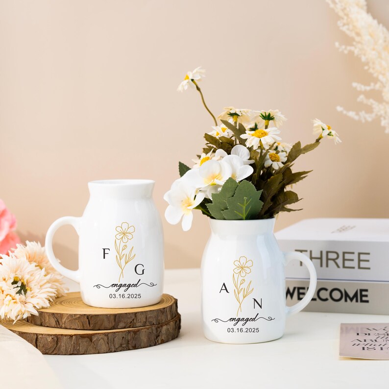 May include: Two white ceramic mugs with gold floral designs and the text "engaged" with the date "03.16.2025". The mugs have the initials "F.G." and "A.N." on them. The mugs are sitting on a wooden surface with a bouquet of white flowers in a vase.
