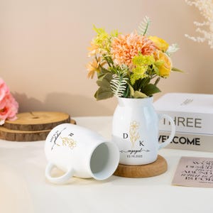 May include: Two white ceramic mugs with gold floral designs and the text "engaged" and "03.16.2025". The mugs are on a wooden surface next to a white ceramic vase with a gold floral design and the text "D K engaged 03.16.2025". The vase is filled with a bouquet of peach and yellow flowers.