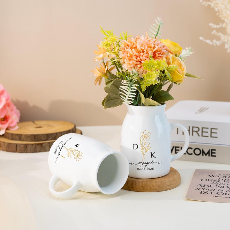 May include: Two white ceramic mugs with gold floral designs and the text "engaged" and "03.16.2025". The mugs are on a wooden surface next to a white ceramic vase with a gold floral design and the text "D K engaged 03.16.2025". The vase is filled with a bouquet of peach and yellow flowers.