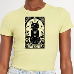 May include: A yellow t-shirt featuring a black and white illustration of a cat within a tarot card style frame. The card reads "THE CAT".