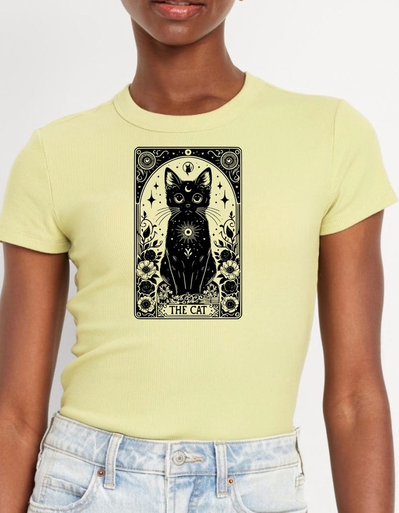 May include: A yellow t-shirt featuring a black and white illustration of a cat within a tarot card style frame. The card reads "THE CAT".