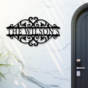 K&ouml;nnte beinhalten: Schwarzes Metallschild mit dem Text "THE WILSON'S" in einer dekorativen Schriftart. Das Schild ist an einer wei&szlig;en Wand mit Marmormuster angebracht, in der N&auml;he einer dunklen Holzt&uuml;r und Gr&uuml;npflanzen.