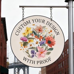 Op de afbeelding: Een rond bord met een bloemenontwerp en de tekst "CUSTOM YOUR DESIGN WITH PROOF". Het bord is crèmekleurig met een zwarte rand en hangt aan een metalen paal met kettingen. Het bloemenontwerp bevat roze, gele en rode bloemen.