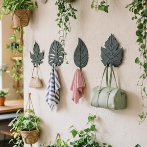 May include: Four dark gray metal wall hooks shaped like various leaves. The hooks hold a small woven bag, a blue and white striped cloth, a red and white striped cloth, and a light green handbag. The hooks are mounted on a textured wall.