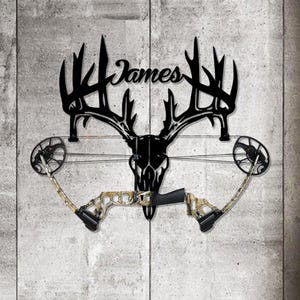 May include: Black metal deer skull with antlers, holding a camouflage compound bow. The name "James" is scripted above the skull. The bow is mounted on a gray, textured wall, creating a rustic, hunting-themed display.