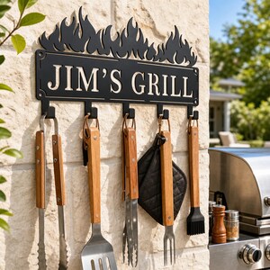 Peut inclure: Un support mural en métal noir avec un bord supérieur en forme de flammes et l'inscription JIM'S GRILL. Le support comporte cinq crochets retenant des ustensiles de barbecue en acier inoxydable avec des manches en bois, dont une pince.