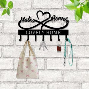 Pode incluir: Porta-chaves de metal preto com os nomes Mateo e Rennie entrelaçados com um coração e as palavras "Lovely Home". O porta-chaves tem seis ganchos e está montado numa parede de tijolo branco.