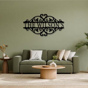 K&ouml;nnte beinhalten: Schwarze Metall-Wandkunst mit dem Text "THE WILSON'S" in einer dekorativen Schriftart. Das Schild hat ein verziertes Rankenmuster um den Text. Das Schild ist an einer gr&uuml;nen Wand &uuml;ber einem Sofa angebracht.