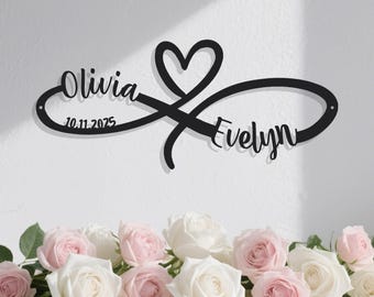 May include: Black metal infinity sign with a heart, featuring the names "Olivia" and "Evelyn" in cursive script. The date "10.11.2025" is also included. The sign is displayed against a white background, with a floral arrangement of pink and white roses at the bottom.