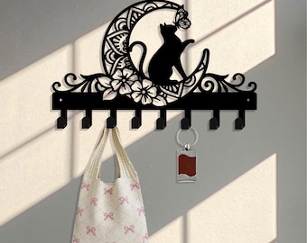 Pode incluir: Porta-chaves de metal preto montado na parede com uma silhueta de gato numa lua crescente com uma borboleta. O porta-chaves tem vários ganchos e é decorado com detalhes florais. Uma sacola branca com laços rosa está pendurada.