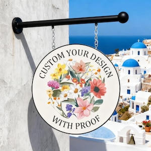 May include: A round, hanging sign with a floral design and the text "CUSTOM YOUR DESIGN WITH PROOF." The sign is suspended by chains from a black metal arm attached to a white wall. The background shows a coastal town with blue domes.