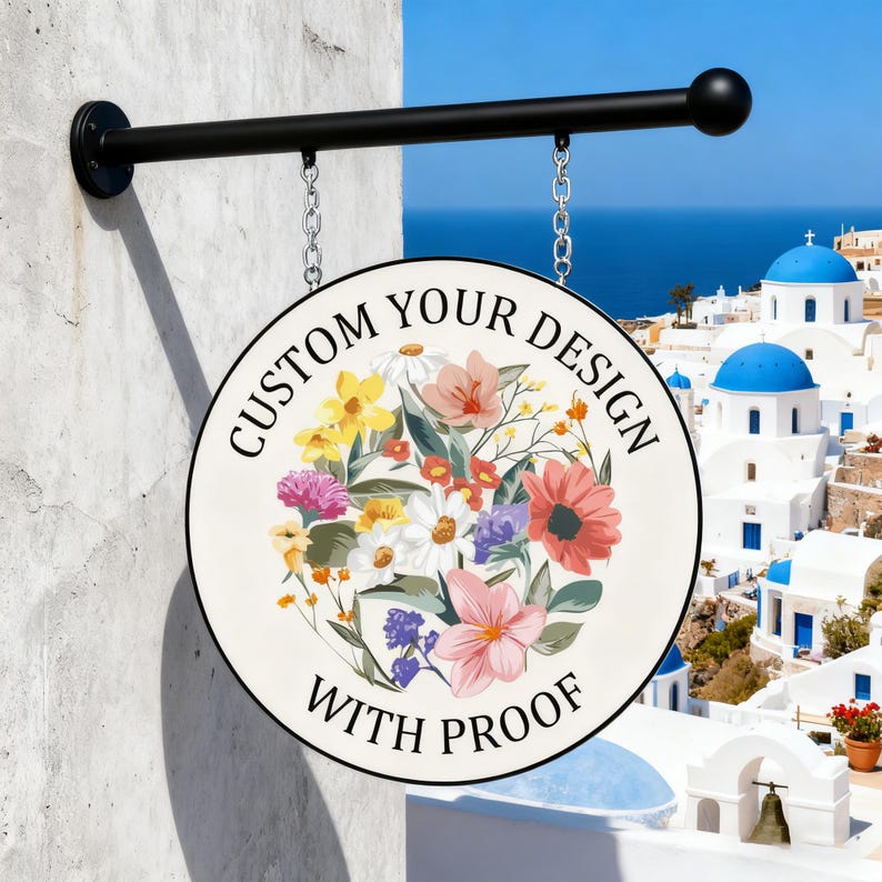 May include: A round, hanging sign with a floral design and the text "CUSTOM YOUR DESIGN WITH PROOF." The sign is suspended by chains from a black metal arm attached to a white wall. The background shows a coastal town with blue domes.