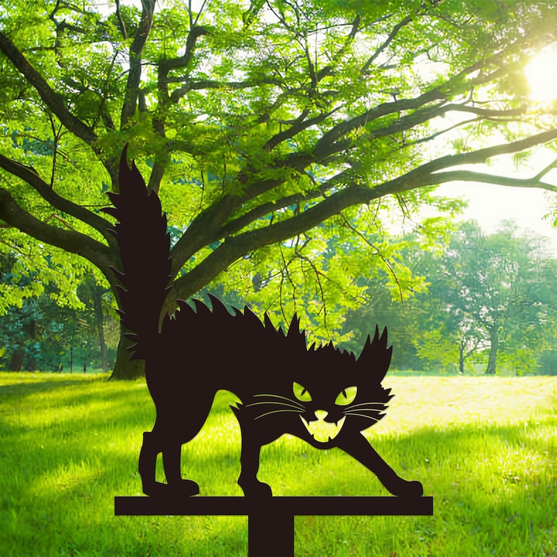 May include: A black metal cat garden stake with a menacing expression, green eyes, and a snarling mouth. The cat has a spiky back and a long, upright tail. The stake is placed in a grassy yard with trees in the background.