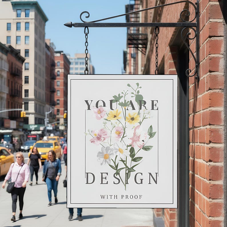 May include: A rectangular sign with the words "YOU ARE DESIGN WITH PROOF" and a floral illustration. The sign is hanging from a black metal bracket on a brick wall, with a city street in the background.