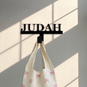 May include: A black metal wall hook with the name "JUDAH" written on it. A cream-colored crocheted tote bag with pink bows hangs from the hook.
