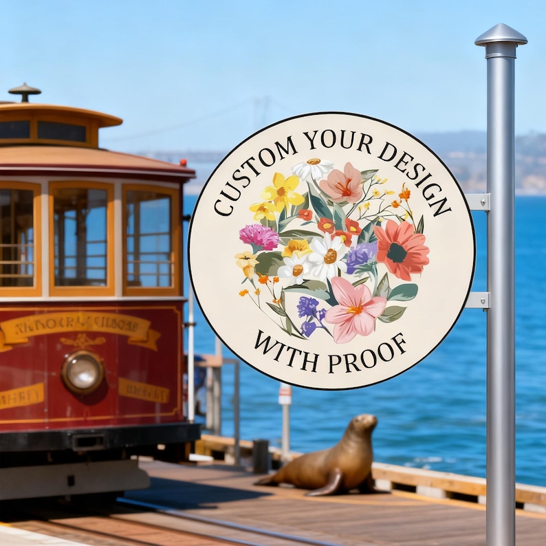 May include: A round sign with the text "CUSTOM YOUR DESIGN WITH PROOF" and a floral design in the center. The sign is mounted on a silver pole. A cable car and a sea lion are in the background.