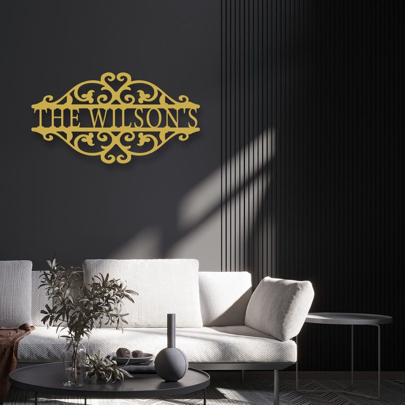 K&ouml;nnte beinhalten: Goldfarbenes Metallschild mit der Aufschrift "THE WILSON'S" in einem verzierten Rahmen. Das Schild ist an einer dunkelgrauen Wand &uuml;ber einem wei&szlig;en Sofa angebracht. Das Design zeigt verschn&ouml;rkelte Verzierungen und einen klassischen, eleganten Stil.