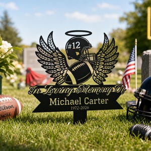 Può includere: Un picchetto commemorativo da giardino in metallo nero con un casco da football americano con il numero 12, un pallone e ali d'angelo. Il testo recita In Loving Memory Of Michael Carter 1972-2026. Include un'aureola sopra il casco.