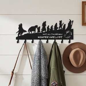 May include: A black metal wall-mounted coat rack featuring a silhouette of a group of travelers and a horse. The design includes the text "NOT ALL THOSE WHO WANDER ARE LOST" and a ring icon. It has seven hooks for hanging items like bags and jackets.