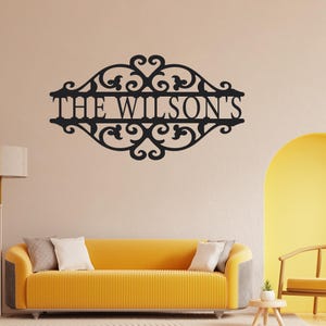K&ouml;nnte beinhalten: Schwarzes Metall-Wandbild mit dem Text "THE WILSON'S" in dekorativer Schrift. Das Schild hat ein verziertes Rankenmuster um den Text. Das Schild ist an einer beigefarbenen Wand &uuml;ber einem gelben Sofa angebracht.