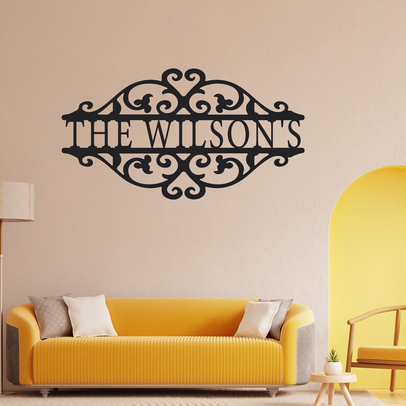 K&ouml;nnte beinhalten: Schwarzes Metall-Wandbild mit dem Text "THE WILSON'S" in dekorativer Schrift. Das Schild hat ein verziertes Rankenmuster um den Text. Das Schild ist an einer beigefarbenen Wand &uuml;ber einem gelben Sofa angebracht.