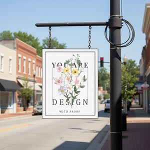 May include: A rectangular sign with a black frame hangs from a black metal bracket. The sign features the words "YOU ARE DESIGN WITH PROOF" around a floral illustration with pink, yellow, and white flowers.