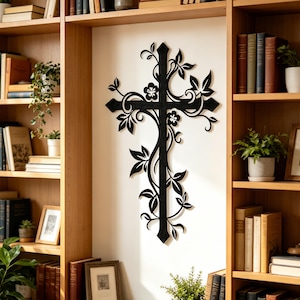May include: A black metal wall art piece shaped like a cross, featuring intricate cut-out designs of swirling vines, leaves, and small five-petaled flowers. The cross has pointed ends and a matte finish, mounted centrally on a plain white wall.