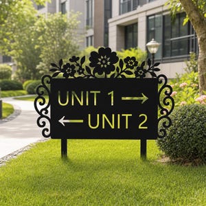 May include: A black metal directional sign featuring a rectangular center with the text UNIT 1 and an arrow pointing right, and UNIT 2 with an arrow pointing left. The sign is decorated with a floral cutout design and scrollwork edges.