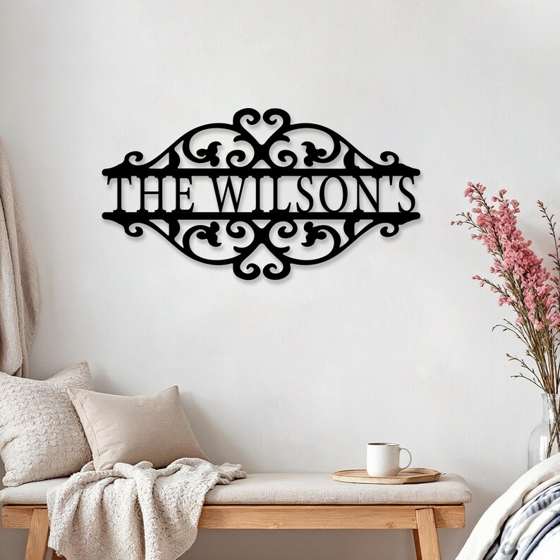 K&ouml;nnte beinhalten: Schwarzes Metallschild mit dem Text "THE WILSON'S" in dekorativer Schrift. Das Schild hat ein verziertes Rankenmuster. Es ist f&uuml;r die Heimdekoration gedacht und an einer wei&szlig;en Wand befestigt.