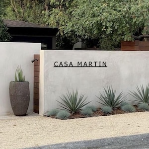 May include: A modern house with a white wall and a black metal sign that reads "CASA MARTIN". The sign is mounted on the wall above a row of green plants.