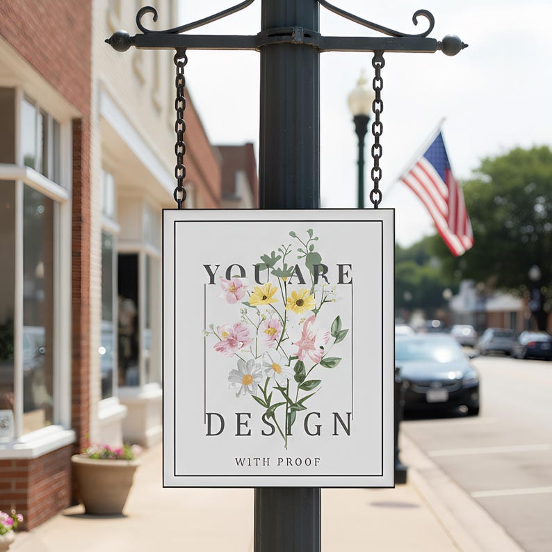 May include: A white sign with a black frame hangs from a black pole. The sign reads "YOU ARE DESIGN WITH PROOF" and features a floral bouquet in pink, yellow, and white. An American flag waves in the background.
