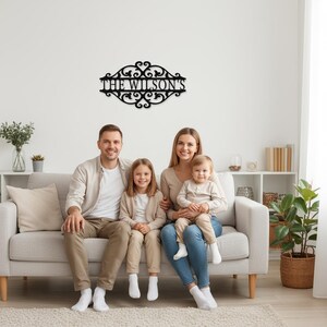 K&ouml;nnte beinhalten: Ein schwarzes Metall-Familiennamenschild mit dem Text "THE WILSON'S" in einer dekorativen Schriftart, montiert an einer wei&szlig;en Wand. Das Schild hat verzierte Schn&ouml;rkel und befindet sich &uuml;ber einer vierk&ouml;pfigen Familie, die auf einem grauen Sofa sitzt.