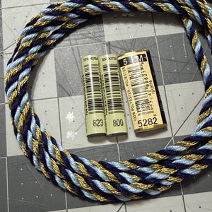 May include: A braided cord in navy blue, light blue, and gold, arranged in a circle. Two spools of thread with the numbers 823 and 800 are visible, along with a label that reads "5 M" and "5282". The items are on a gray grid surface.