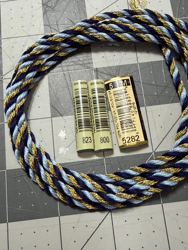 May include: A braided cord in navy blue, light blue, and gold, arranged in a circle. Two spools of thread with the numbers 823 and 800 are visible, along with a label that reads "5 M" and "5282". The items are on a gray grid surface.