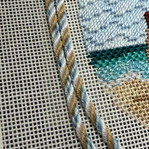May include: Close-up of needlepoint with twisted cord detail. The cord is a mix of white, light blue, and gold. The background shows a beach scene with blue sky and green water. The needlepoint is on a white mesh canvas.