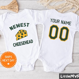 May include: Two white baby onesies. One features the text "NEWEST CHEESEHEAD" with a cheese graphic. The other has "YOUR NAME" and the number "00" in green and gold. These onesies are designed for babies.