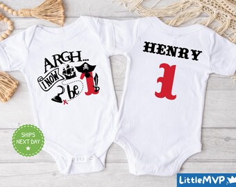 Può includere: Body per neonati bianco con un design a tema pirata. La parte anteriore presenta il testo "Argh... I now be" con una nave pirata, un teschio e ossa incrociate. Il retro ha il nome "HENRY" e il numero "1" in rosso.