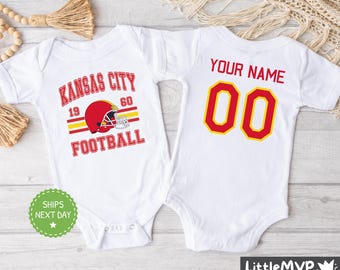May include: Two white baby onesies. One onesie has the text "Kansas City Football" with a red football helmet graphic. The other onesie has the text "Your Name 00".