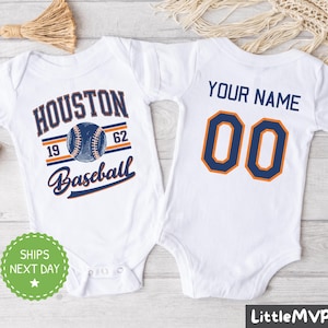 May include: White baby onesie with a blue and orange baseball design. The front of the onesie says "Houston Baseball" with a baseball graphic and the numbers "19" and "62". The back of the onesie says "Your Name" with the number "00" in orange and blue.