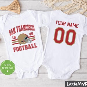 Peut inclure: Deux bodies blancs pour bébé. Un body a le texte "San Francisco Football" avec un graphique de casque de football. L'autre body a le texte "Your Name 00".