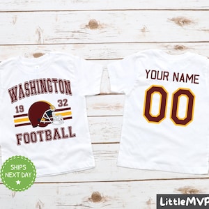 May include: White t-shirt with a burgundy and gold football design on the front. The design includes the words "Washington Football" and a football helmet. The back of the shirt has a burgundy and gold number "00" with the text "Your Name" above it.