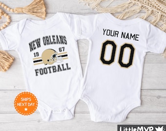 Puede incluir: Dos bodies blancos para bebé. Un body tiene un gráfico de casco de fútbol de los New Orleans Saints en negro y dorado con el texto "New Orleans Football" y los números "19" y "67". El otro body tiene el texto "Your Name" y el número "00" en negro.