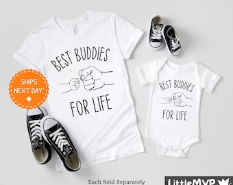 Puede incluir: Dos camisetas blancas con el texto "Best Buddies for Life" y un gráfico de dos puños chocando. La camiseta más grande tiene manga corta y la más pequeña es un body.