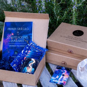 May include: A brown box with a blue and gold deck of cards, a book titled "Primer Oraculo de Constelaciones Familiares", and a white crystal. The box is open and the cards are visible. The book is titled "Primer Oraculo de Constelaciones Familiares" and has the text "Sanando tus raices" on the cover.